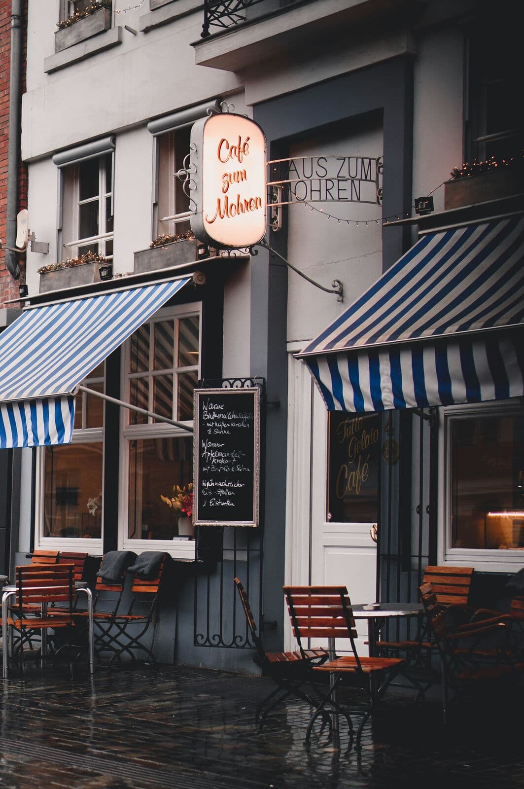 Gemütliches Café mit gestreiften Markisen, leuchtendem Schild und Stühlen vor der Tür