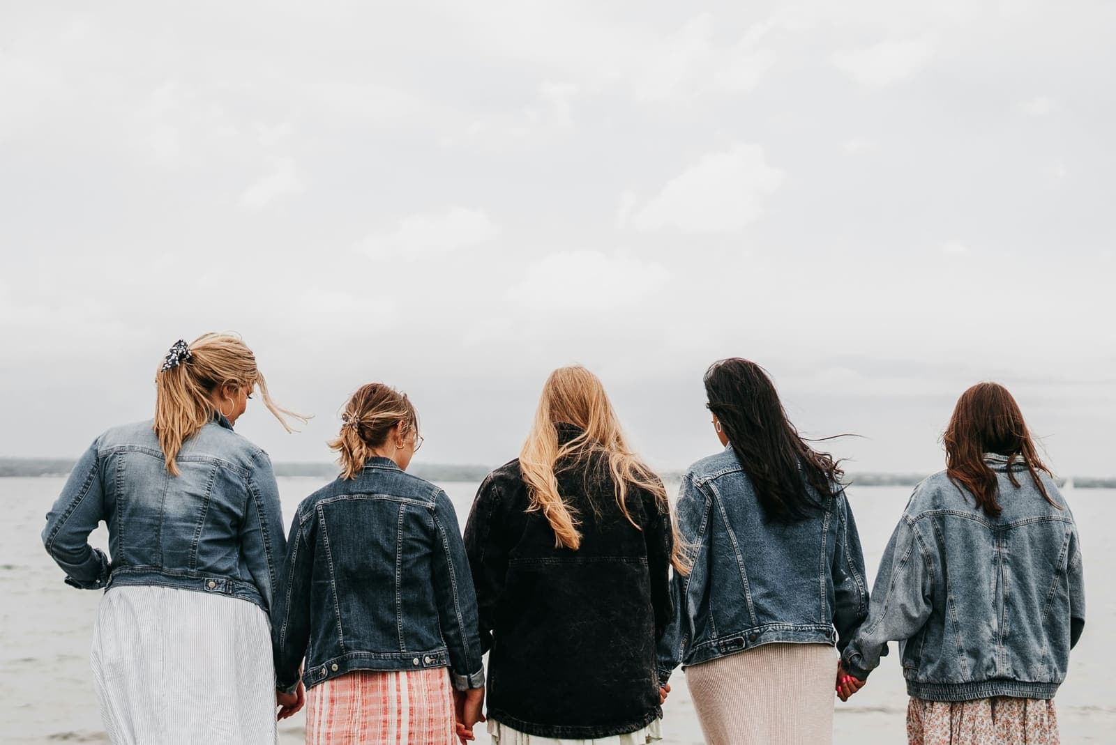 Fünf Frauen in Jeansjacken halten sich an den Händen und blicken aufs Wasser